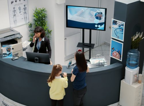 Receptionist working in hospital waiting area and offering support to patients at checkup visit, mother with child having medical appointment with physician. Reception desk at healthcare clinic.